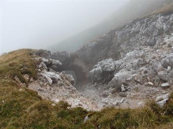 Transitando sotto il pendio  occidentale di un'anticima, il sentiero si fà ora più incerto, percorrendo un'esile crestina erbosa caratterizzata da un grande foro naturale in basso a sinistra.<br />